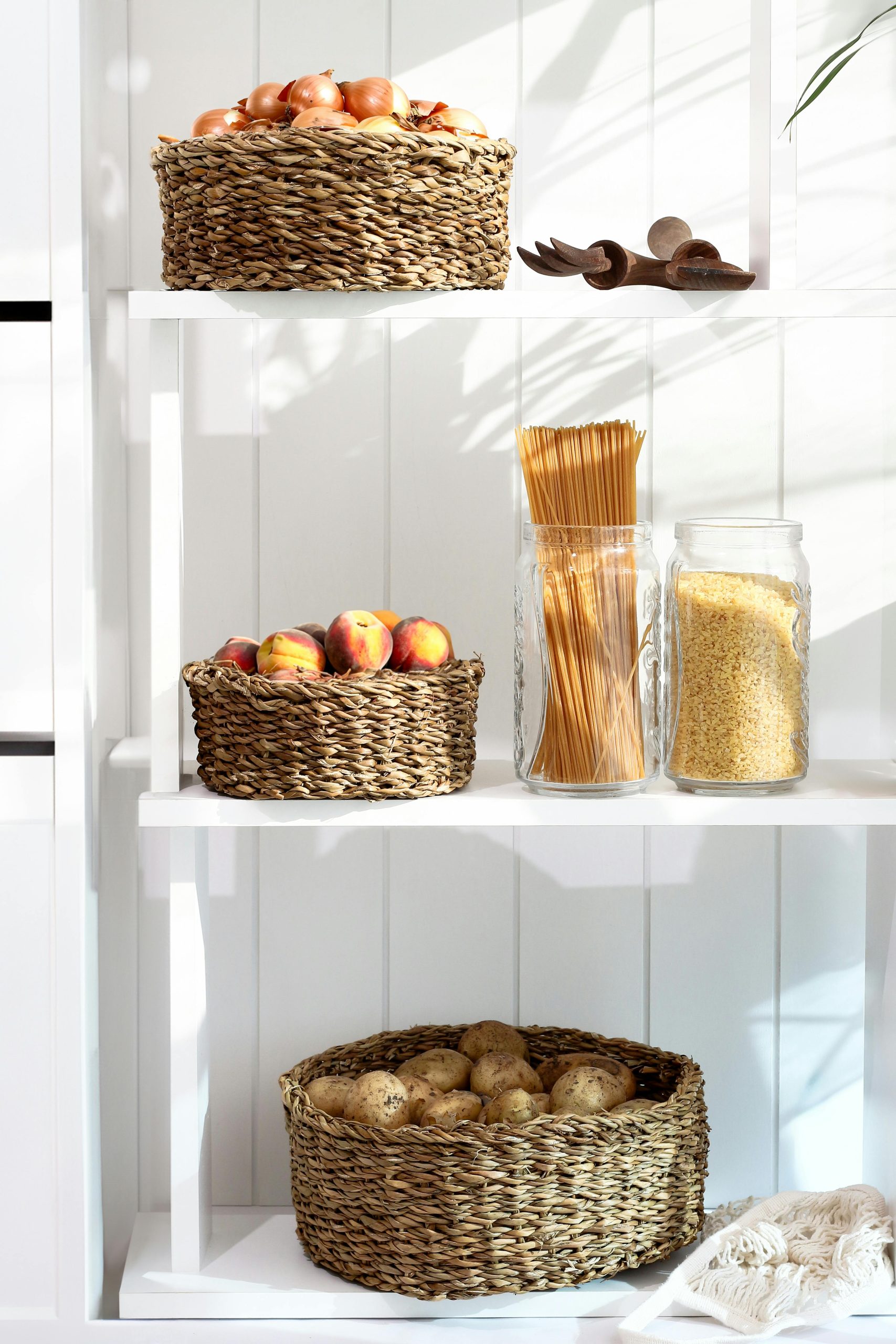 A neatly organized pantry shelf with wicker baskets holding apples, peaches, and potatoes, alongside pasta and grains.